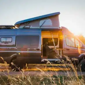Tree House Sprinter 4x4 Campervan mit geöffnetem Aufstelldach und geöffneter Seitentür auf einem Feldweg bei Sonnenuntergang