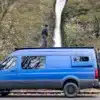 Blauer Mercedes Sprinter 4x4 Campervan mit Dachträger und Fahrradträger, geparkt vor einem Wasserfall, Person steht auf dem Dach