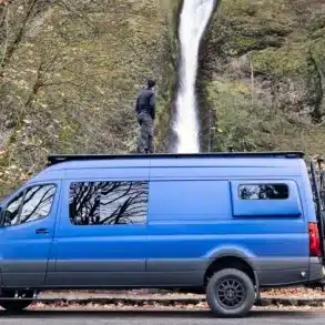 Blauer Mercedes Sprinter 4x4 Campervan mit Dachträger und Fahrradträger, geparkt vor einem Wasserfall, Person steht auf dem Dach