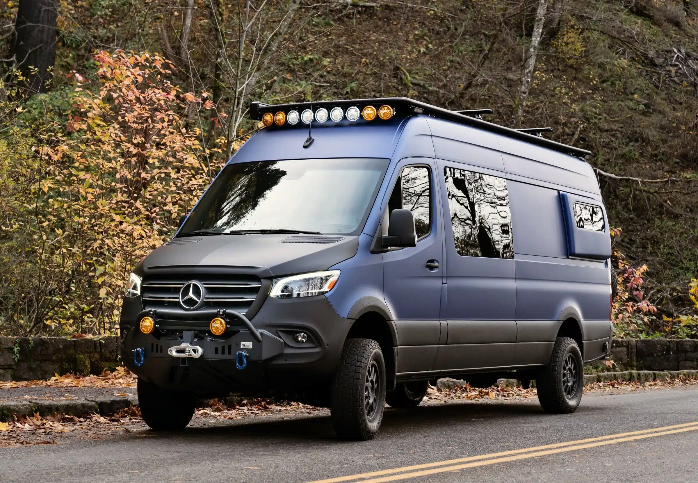 Navyblauer Mercedes Sprinter 4x4 Campervan mit Offroad-Umbau und Dachträger im herbstlichen Wald