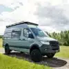 Ein ausgebauter 2019 Mercedes Sprinter 4x4 Walker steht auf einem Feldweg, umgeben von grüner Wiese und Bäumen, unter bewölktem Himmel.