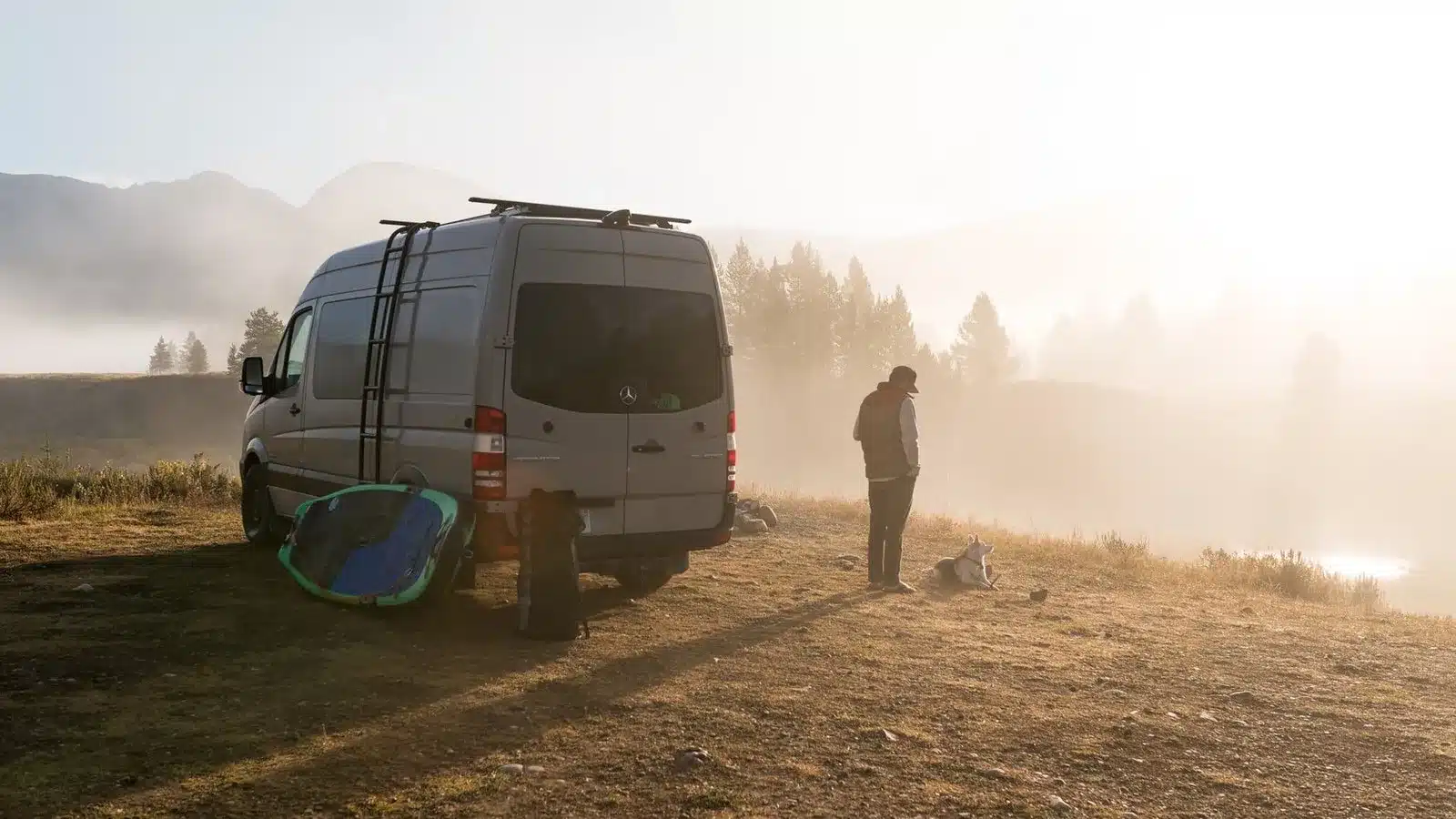 Campervan und Mensch mit Hund auf einer Wiese im Morgennebel vor bewaldeten Bergen