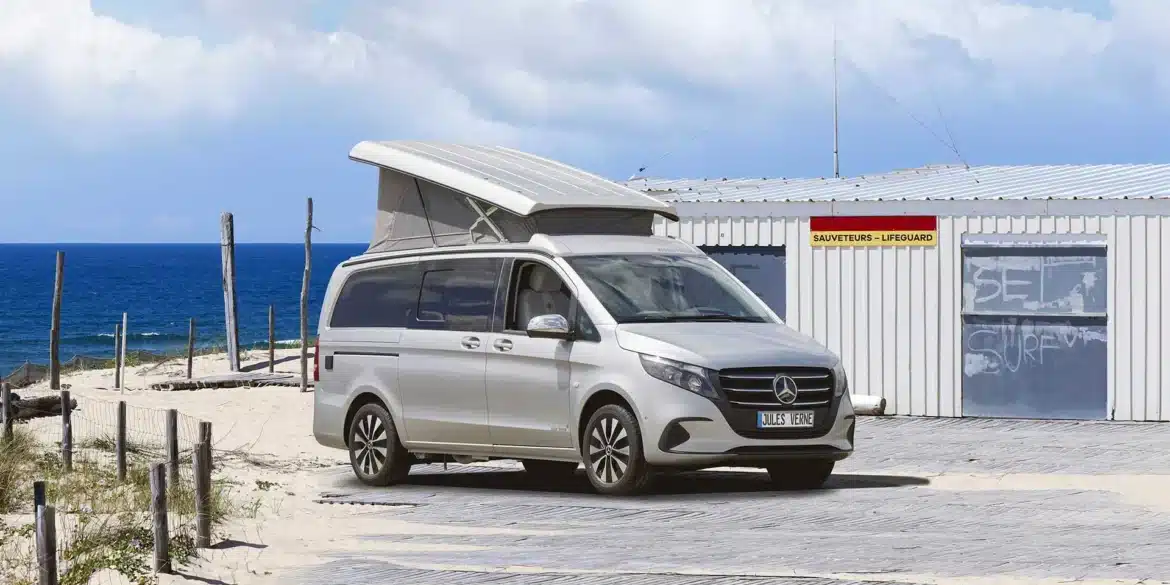 Westfalia Jules Verne Campervan mit geöffnetem Aufstelldach am Strand, im Hintergrund ein Rettungshäuschen und das Meer