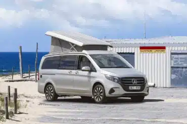 Westfalia Jules Verne Campervan mit geöffnetem Aufstelldach am Strand, im Hintergrund ein Rettungshäuschen und das Meer