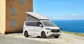 Weißer Westfalia Kelsey Campervan mit geöffnetem Aufstelldach steht vor einem Haus mit Blick auf das Meer