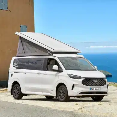 Weißer Westfalia Kelsey Campervan mit geöffnetem Aufstelldach steht vor einem Haus mit Blick auf das Meer