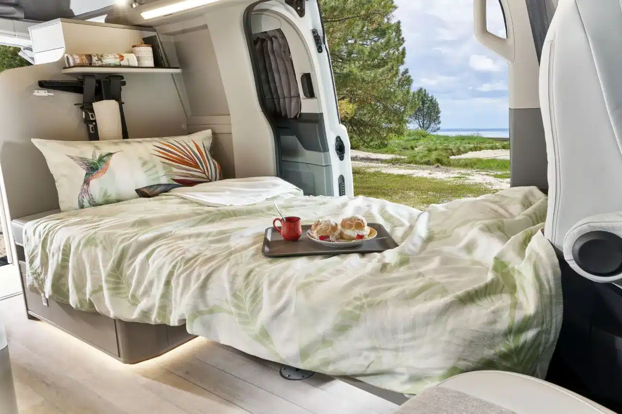 Innenansicht eines Westfalia Kelsey Campervans mit ausgeklapptem Bett, Tablett mit Frühstück und Blick ins Grüne durch die geöffnete Schiebetür