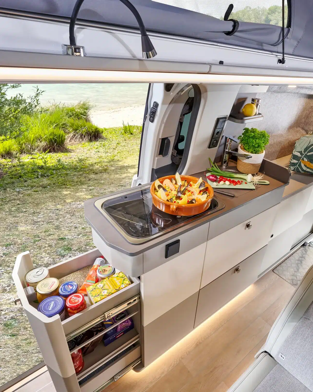 Innenansicht der Camper-Küche im Westfalia Kipling mit ausziehbarem Vorratsschrank, Arbeitsfläche, Kräutern und Seeblick