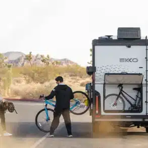 Winnebago EKKO Wohnmobil mit geöffneter Heckgarage, in der Fahrräder verstaut sind, zwei Personen im Vordergrund, Wüstenlandschaft im Hintergrund