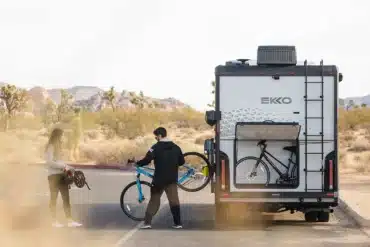 Winnebago EKKO Wohnmobil mit geöffneter Heckgarage, in der Fahrräder verstaut sind, zwei Personen im Vordergrund, Wüstenlandschaft im Hintergrund