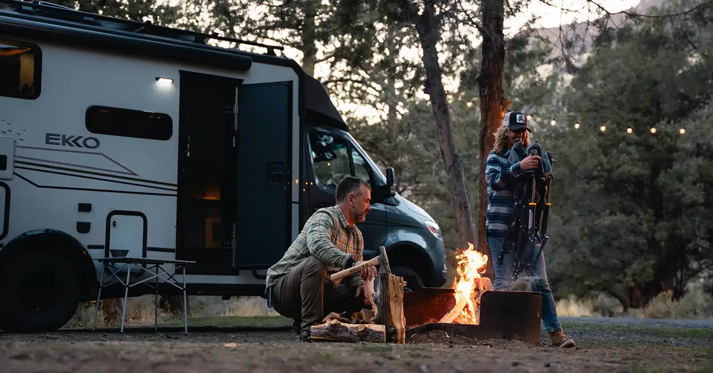 Winnebago EKKO Wohnmobil im Wald, zwei Personen am Lagerfeuer vor dem Fahrzeug