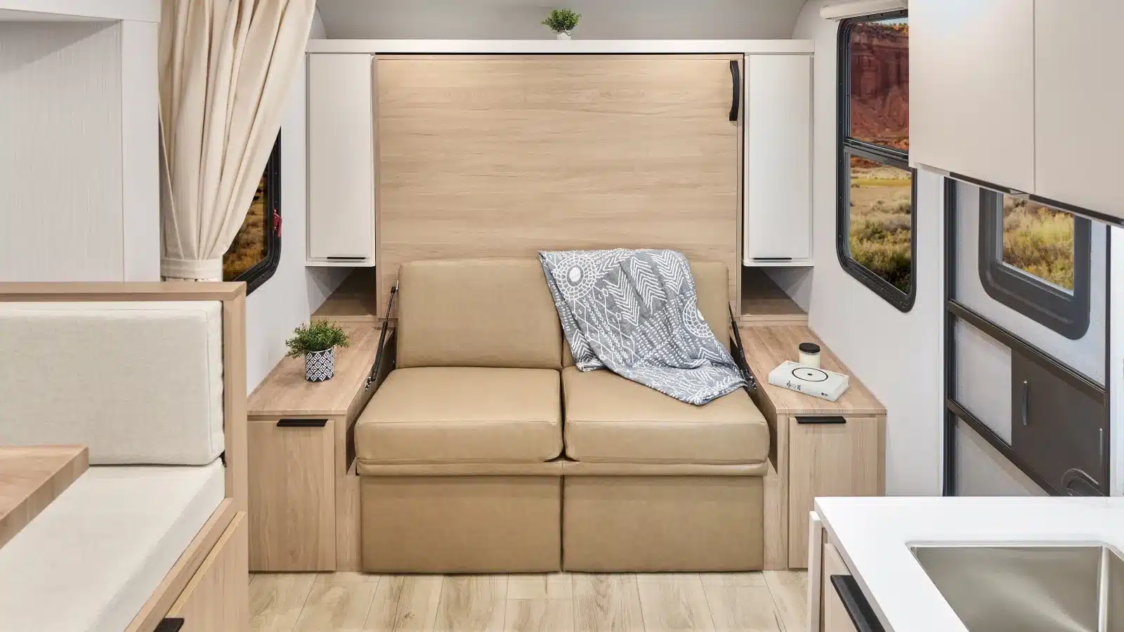 Wohnbereich eines Winnebago Micro Minnie mit beigem Sofa, Murphy-Bett, hellen Holzmöbeln und großen Fenstern