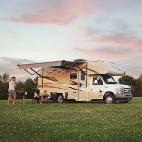Winnebago Minnie Winnie NPF Wohnmobil mit ausgefahrener Markise, Familie beim Camping auf einer Wiese