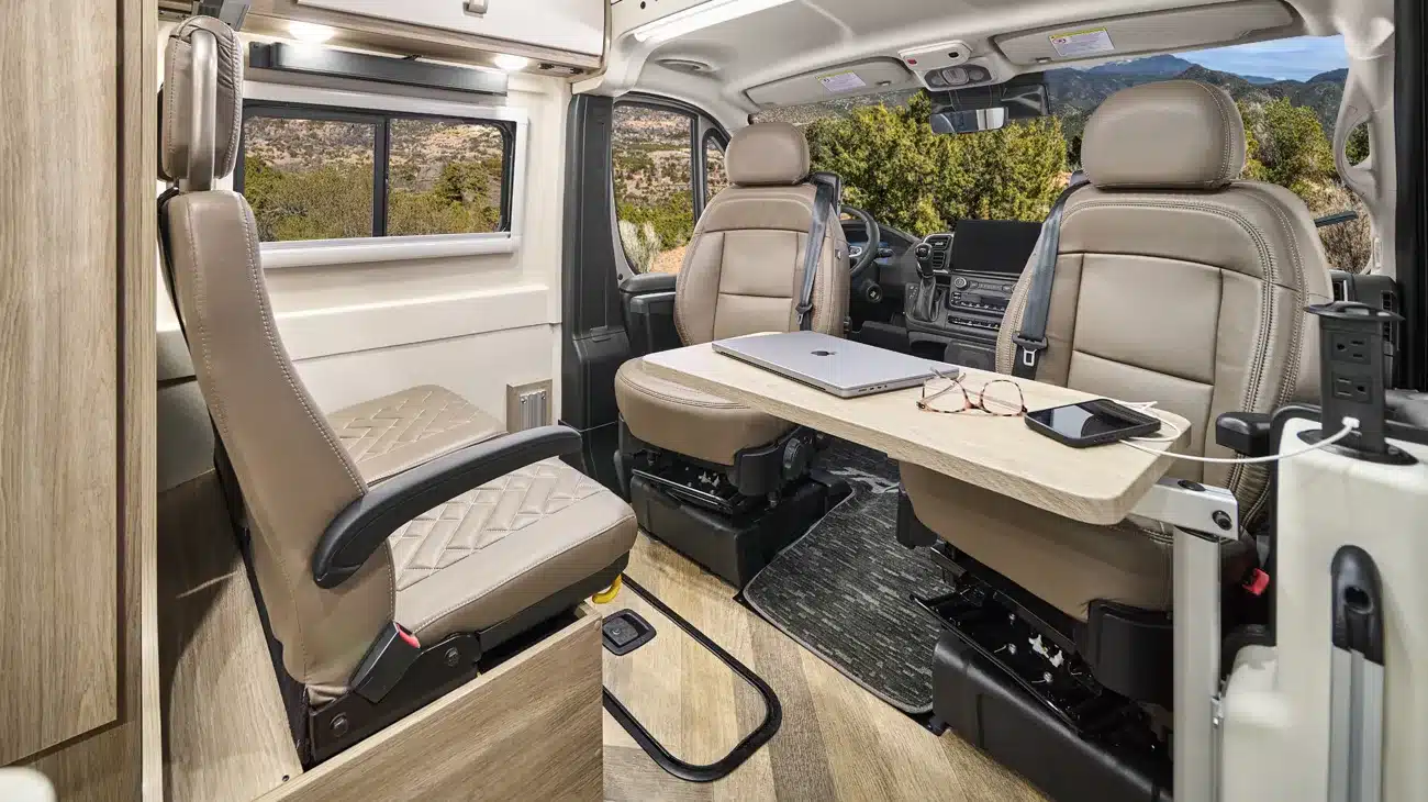 Innenansicht des Winnebago Travato mit drehbaren Vordersitzen, Tisch, Laptop und Ausblick durch das Fenster