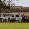 Winnebago Voyage Travel Trailer mit ausgefahrener Markise, Campingstühlen und Zugfahrzeug auf einer Wiese vor herbstlicher Waldkulisse