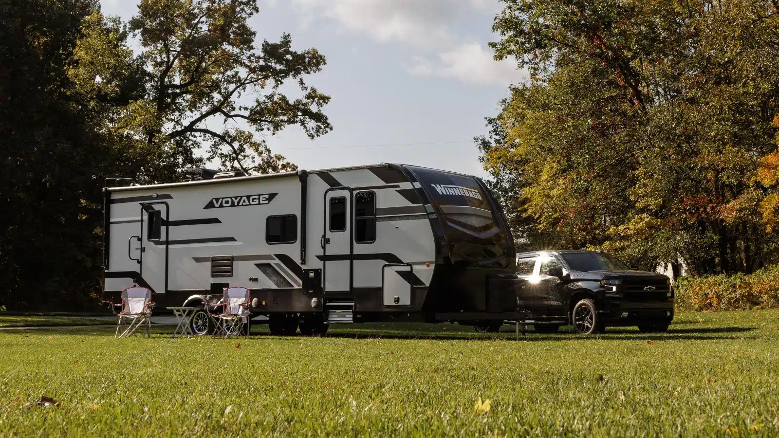 Winnebago Voyage Travel Trailer mit ausgefahrener Markise, Campingstühlen und Zugfahrzeug auf einer Wiese