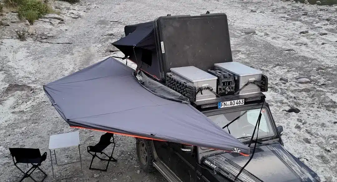Fahrzeug mit 180-Grad-Markise, Campingtisch und Stühlen auf steinigem Gelände am Flussufer.