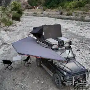 Fahrzeug mit 180-Grad-Markise, Campingtisch und Stühlen auf steinigem Gelände am Flussufer.