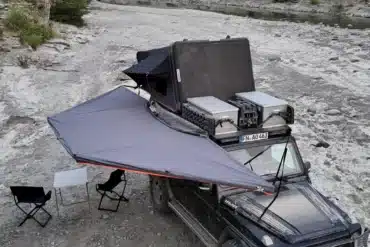 Fahrzeug mit 180-Grad-Markise, Campingtisch und Stühlen auf steinigem Gelände am Flussufer.