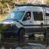 Storyteller Overland Classic MODE XO Allrad-Campervan fährt durch ein flaches Gewässer im Wald.