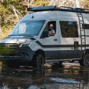 Storyteller Overland Classic MODE XO Allrad-Campervan fährt durch ein flaches Gewässer im Wald.