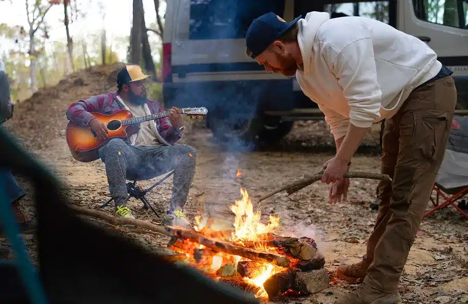 Outdoor-Szene mit zwei Männern am Lagerfeuer und geparktem Campervan im Hintergrund