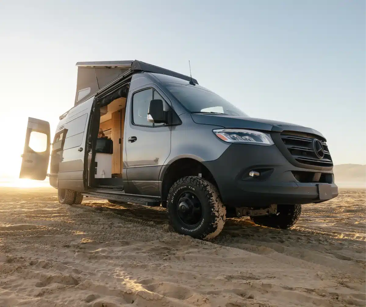2026 Crew Mode XL Campervan auf Basis Mercedes-Benz Sprinter, mit aufgeklapptem LunarLoft-Dach am Strand, Allrad und Offroad-Bereifung, Schiebetür geöffnet.
