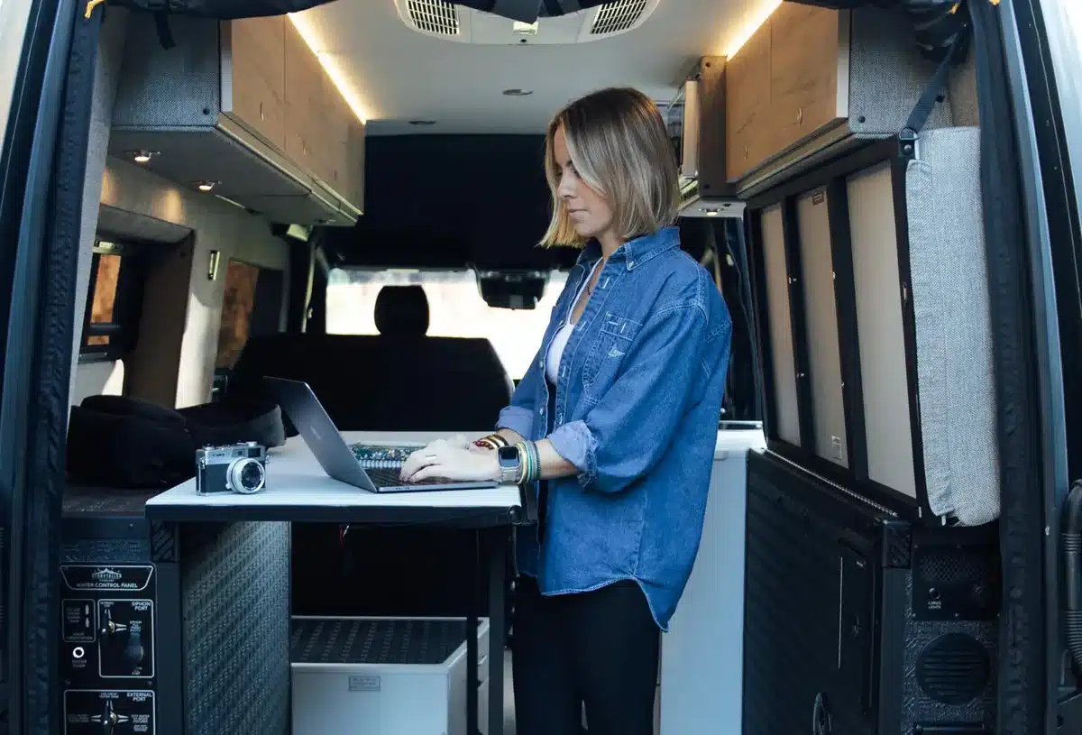 Frau arbeitet mit Laptop im Innenraum eines 2026 Dark MODE OG Kastenwagens, umgeben von Schränken und Technik.