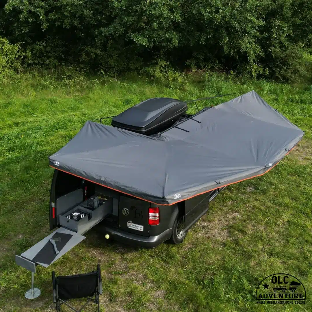 270 Grad freitragende Markise Xtended an einem Campingbus mit ausgezogener Heckküche und Campingausstattung auf einer Wiese.