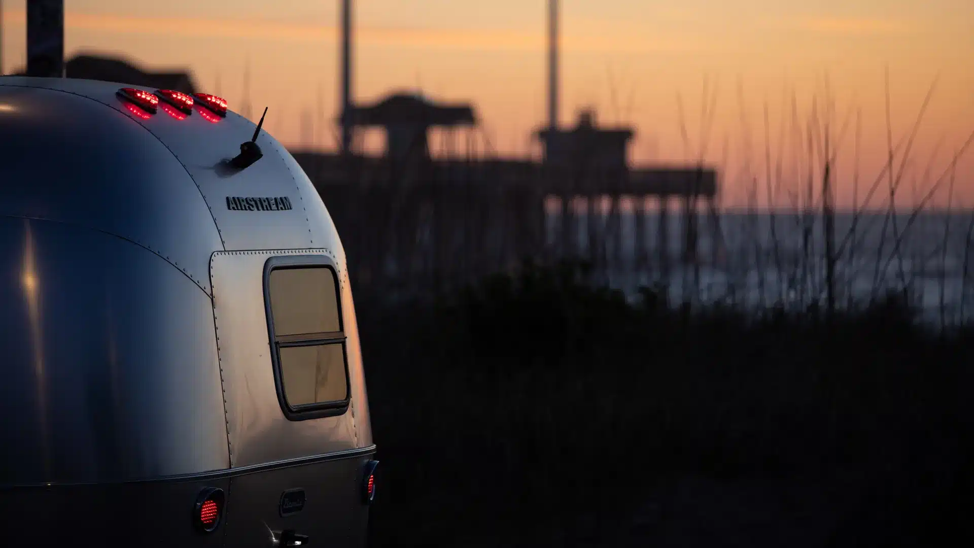 Airstream Bambi 16RB Caravan von hinten am Abend, mit leuchtenden Rücklichtern und Fenster, im Hintergrund eine Küstenlandschaft bei Sonnenuntergang