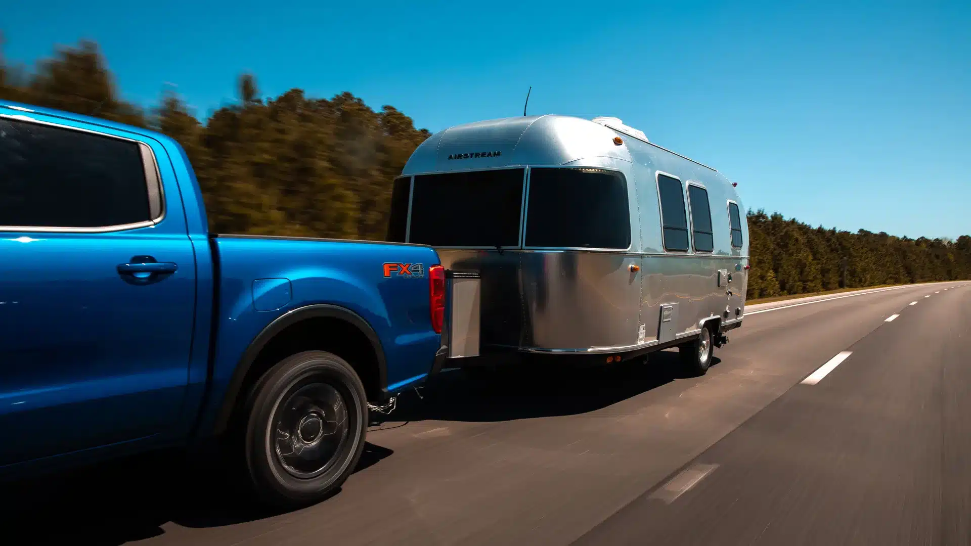 Airstream Bambi 20FB Wohnwagen, gezogen von einem blauen Pickup-Truck auf einer Landstraße bei Sonnenschein.