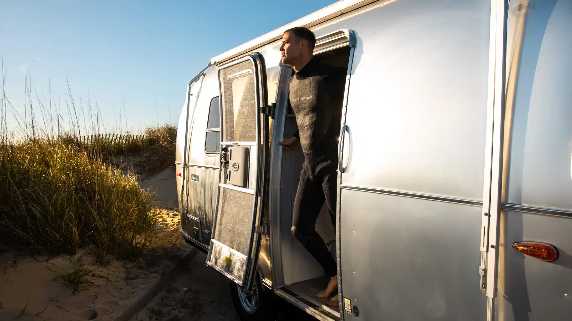 Ein Mann steht in der Tür eines silbernen Airstream Bambi 20FB Wohnwagens, der an einem naturbelassenen Sandstrand mit Dünen parkt.