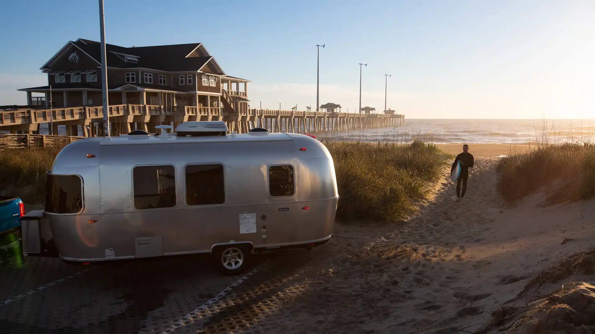 Airstream Bambi 20FB Wohnwagen am Meer neben einem Steg, ein Surfer läuft Richtung Strand