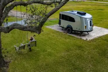 Airstream Basecamp 20Xe Wohnwagen auf einer Rasenfläche mit Baumschaukel, isoliert in der Natur, Seitenansicht.