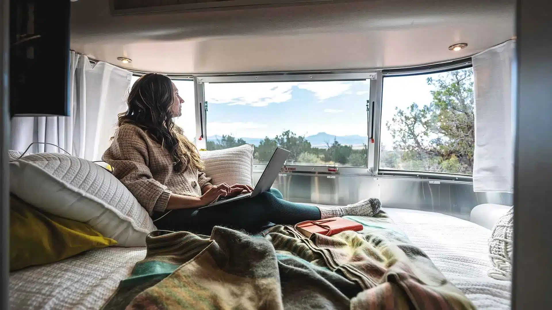 Frau arbeitet am Laptop auf dem Heckbett im Airstream Caravel 16RB mit Panoramafenster und Ausblick auf die Natur.