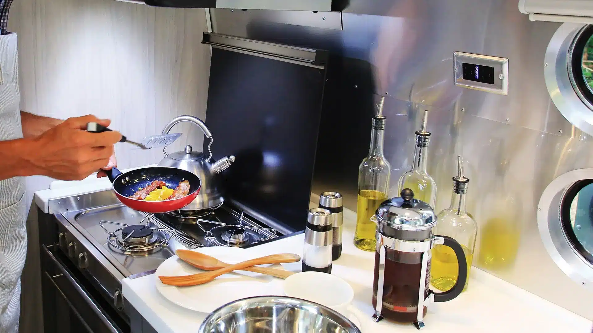 Küchenbereich eines Airstream Globetrotter Wohnanhängers mit Kochfeld, Küchenutensilien und Person beim Frühstück zubereiten.