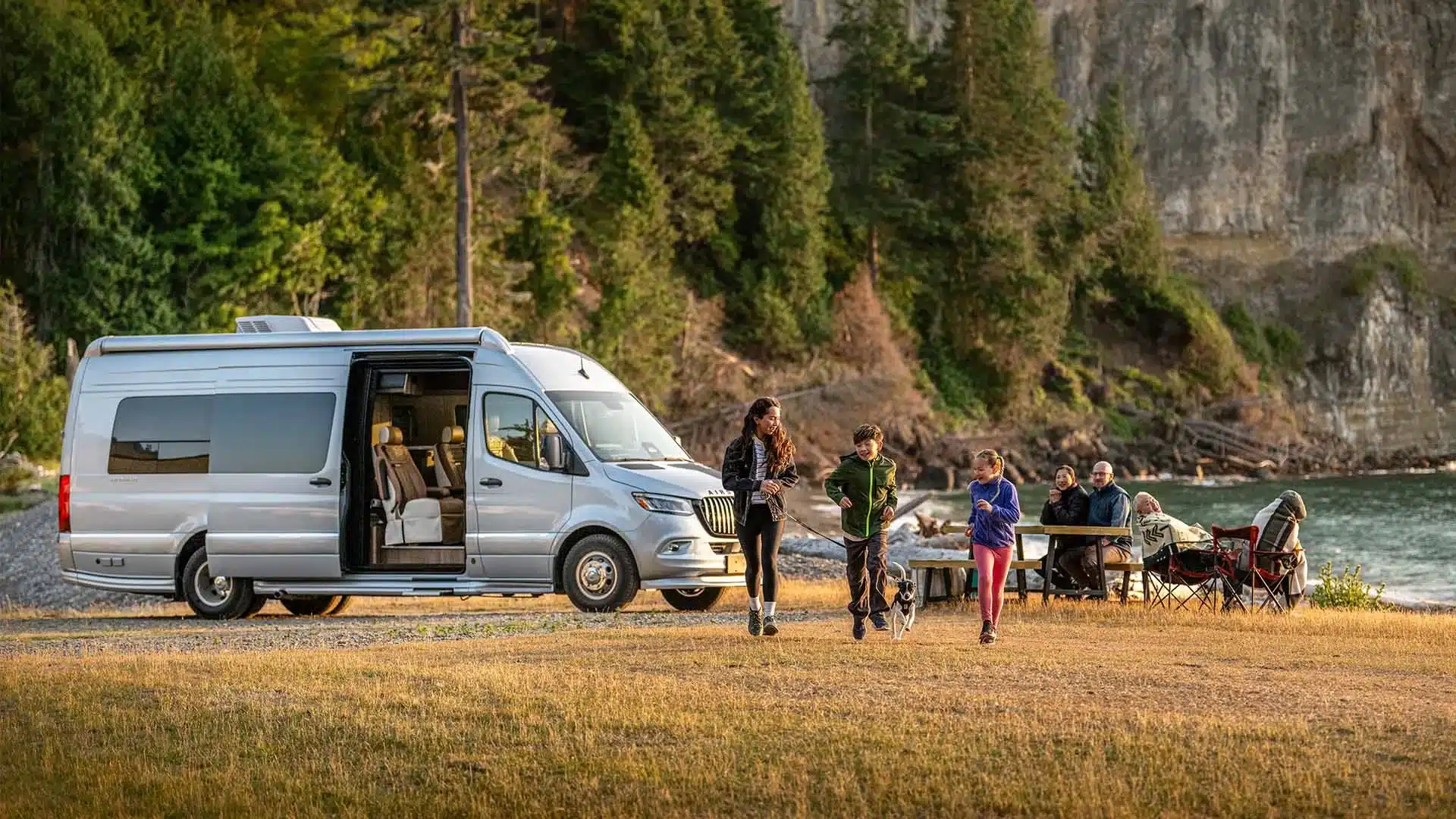 Airstream Interstate 24GL Campervan steht geöffnet an einem Seeufer, Menschen sitzen und spielen in der Natur.