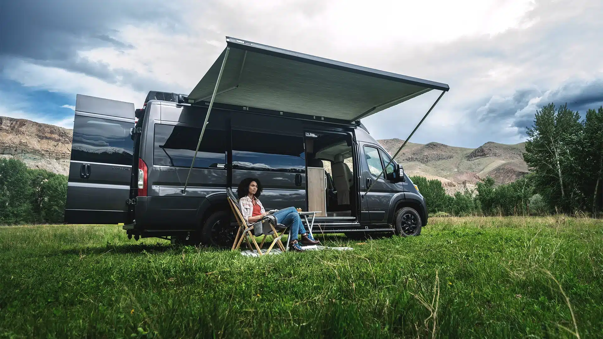 Airstream Rangeline 2026 Kastenwagen mit Markise und Camperin auf Campingstuhl in der Natur.
