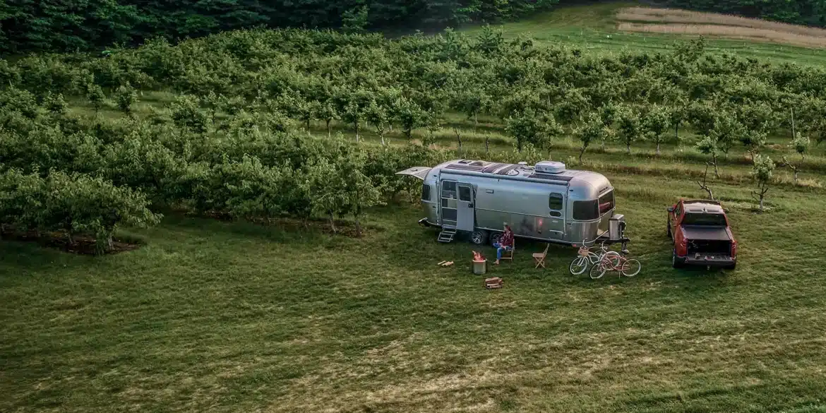 Airstream Trade Wind 2026 Reiseanhänger mit Pickup auf einer grünen Wiese, umgeben von Obstbäumen.