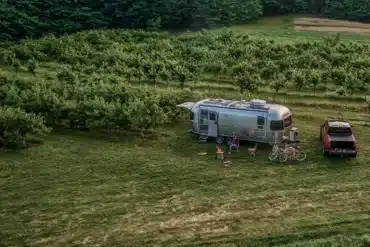 Airstream Trade Wind 2026 Reiseanhänger mit Pickup auf einer grünen Wiese, umgeben von Obstbäumen.