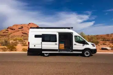 Seitenansicht eines weißen Ford Transit Extended Campervans mit Dachträger und Markise, geparkt vor einer Wüstenlandschaft unter blauem Himmel
