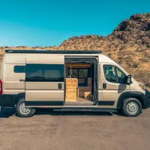 RAM Promaster 2500 High Roof 159 mit geöffnetem Seiteneinstieg, Blick auf maßgefertigten Innenausbau aus Holz in karger Wüstenlandschaft