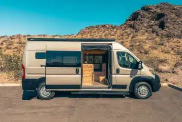 RAM Promaster 2500 High Roof 159 mit geöffnetem Seiteneinstieg, Blick auf maßgefertigten Innenausbau aus Holz in karger Wüstenlandschaft