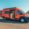 Seitenansicht eines orangefarbenen Blazestar Campervans mit geöffneter Seitentür und sichtbarem Innenausbau auf einer Straße in sonniger Landschaft.