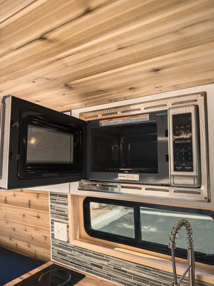 Offene Mikrowelle im Küchenbereich eines ausgebauten Camper Vans mit Holzdecke und Fenster