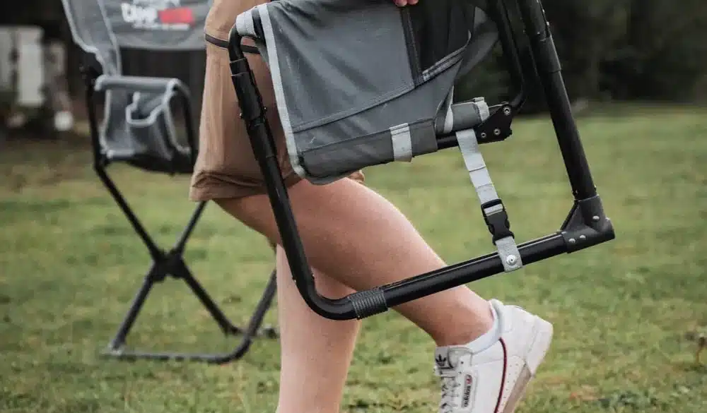 Person trägt einen zusammengefalteten CAMPWERK Campingstuhl über eine Wiese, im Hintergrund steht ein aufgebauter Stuhl.