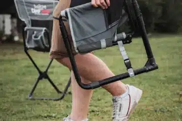 Person trägt einen zusammengefalteten CAMPWERK Campingstuhl über eine Wiese, im Hintergrund steht ein aufgebauter Stuhl.