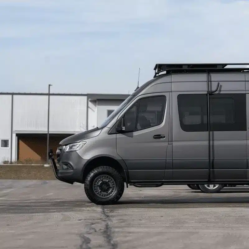 Grauer Cape Breton Campervan von Vancini Conversions auf einem Parkplatz, Seitenansicht mit Offroad-Bereifung und Dachträger.
