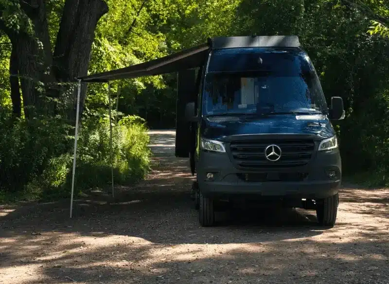 Der Copper Falls Campervan von VanCini Conversions steht mit ausgefahrener Markise im Grünen auf einem Waldweg.