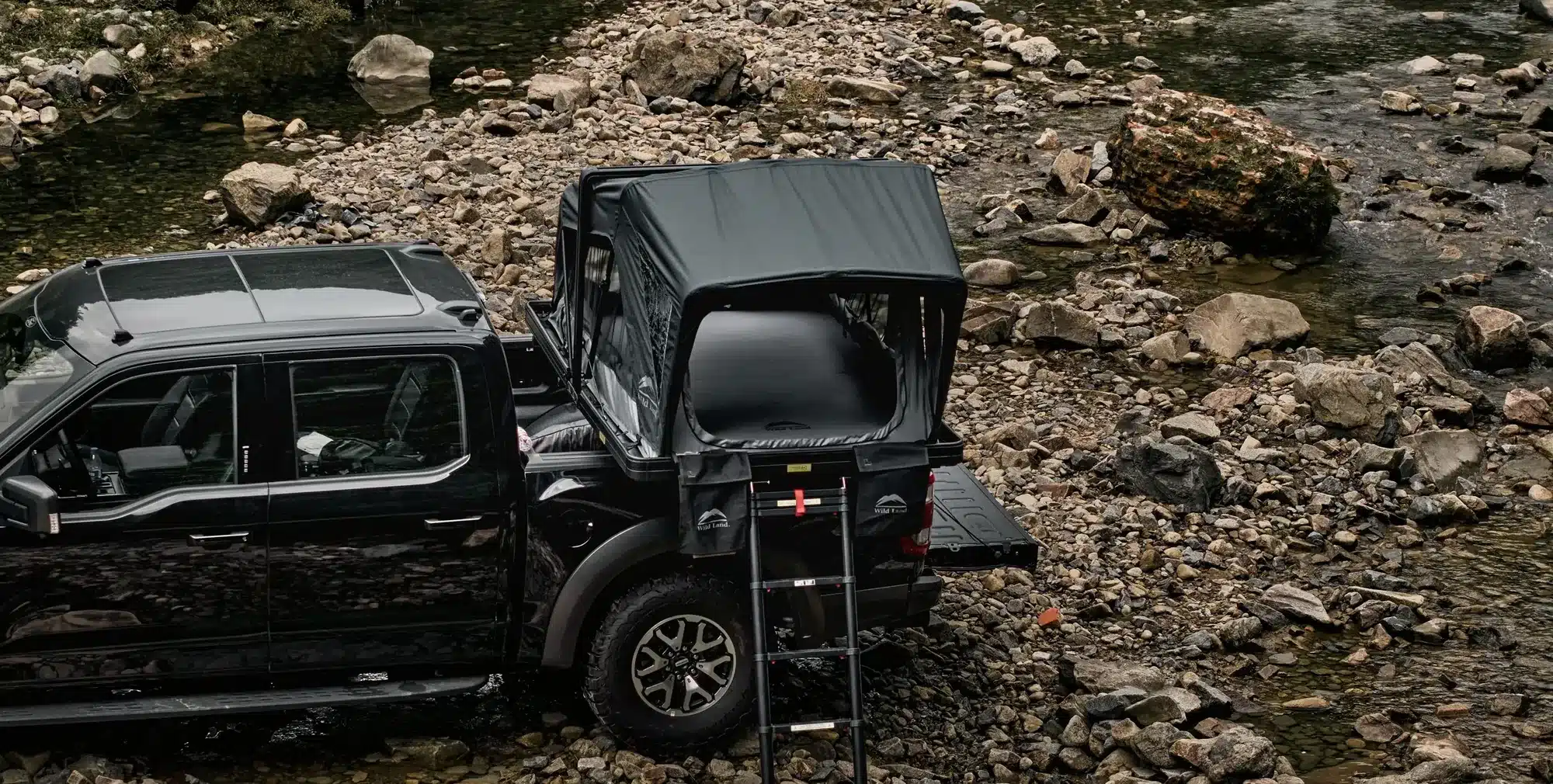 Hartschalen-Dachzelt Summit Explorer 140 von Wild Land International auf Pick-up, aufgestellt an einem steinigen Flussufer mit montierter Leiter.